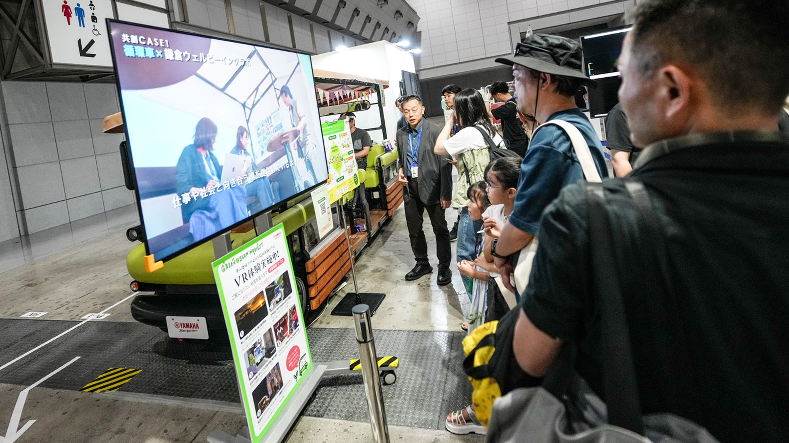 大型モニターとグリーンの車両を展示したブースで、スタッフが来場者に説明する様子