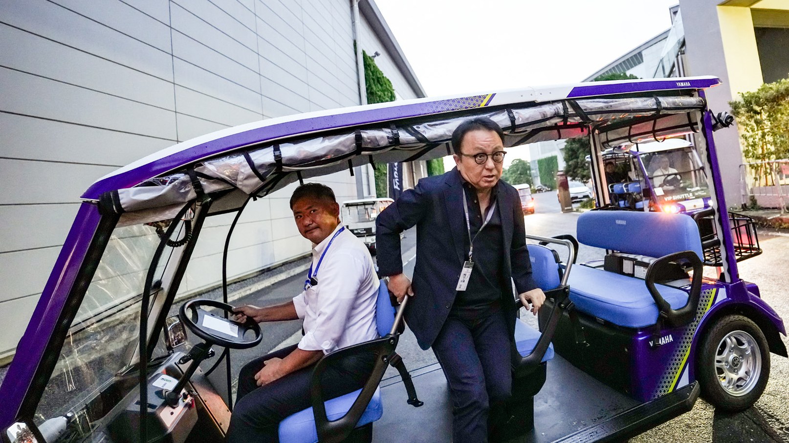 紫色のグリスロの座席から降りようとするスーツ姿の男性と、運転席に座るスタッフ
